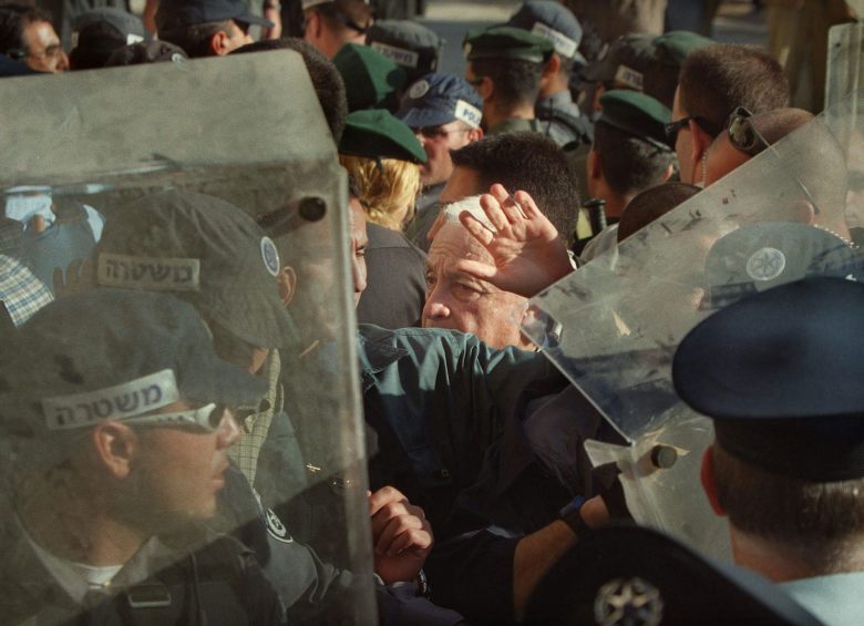 A man rises his hand as he is surrounded by security guards some with shields.